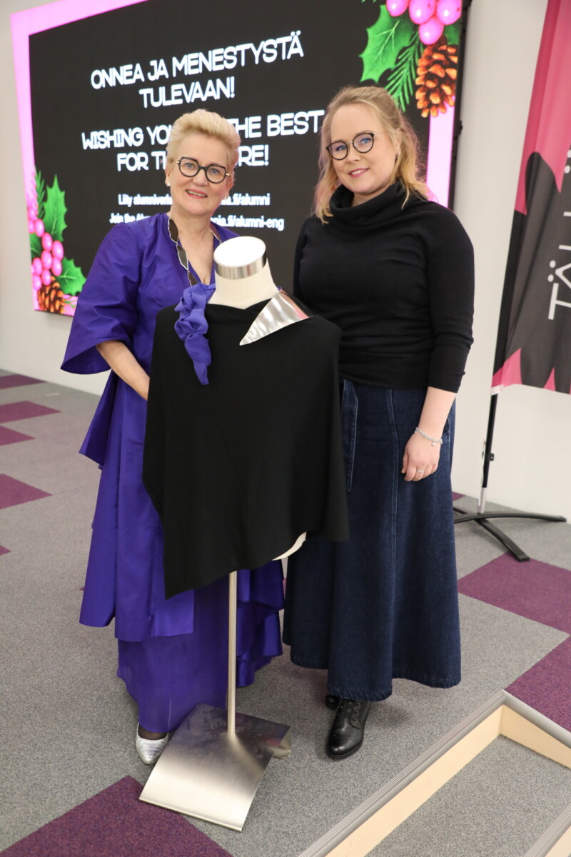 Kaksi naista seisoo hymyillen mallinukessa olevan mustan vaatteen vieressä. Toisella naisella on violetti mekko, toisella musta yläosa ja pitkä sininen hame. Taustalla on suuri näyttö, jossa on onnittelutekstiä ja juhlallista grafiikkaa.
