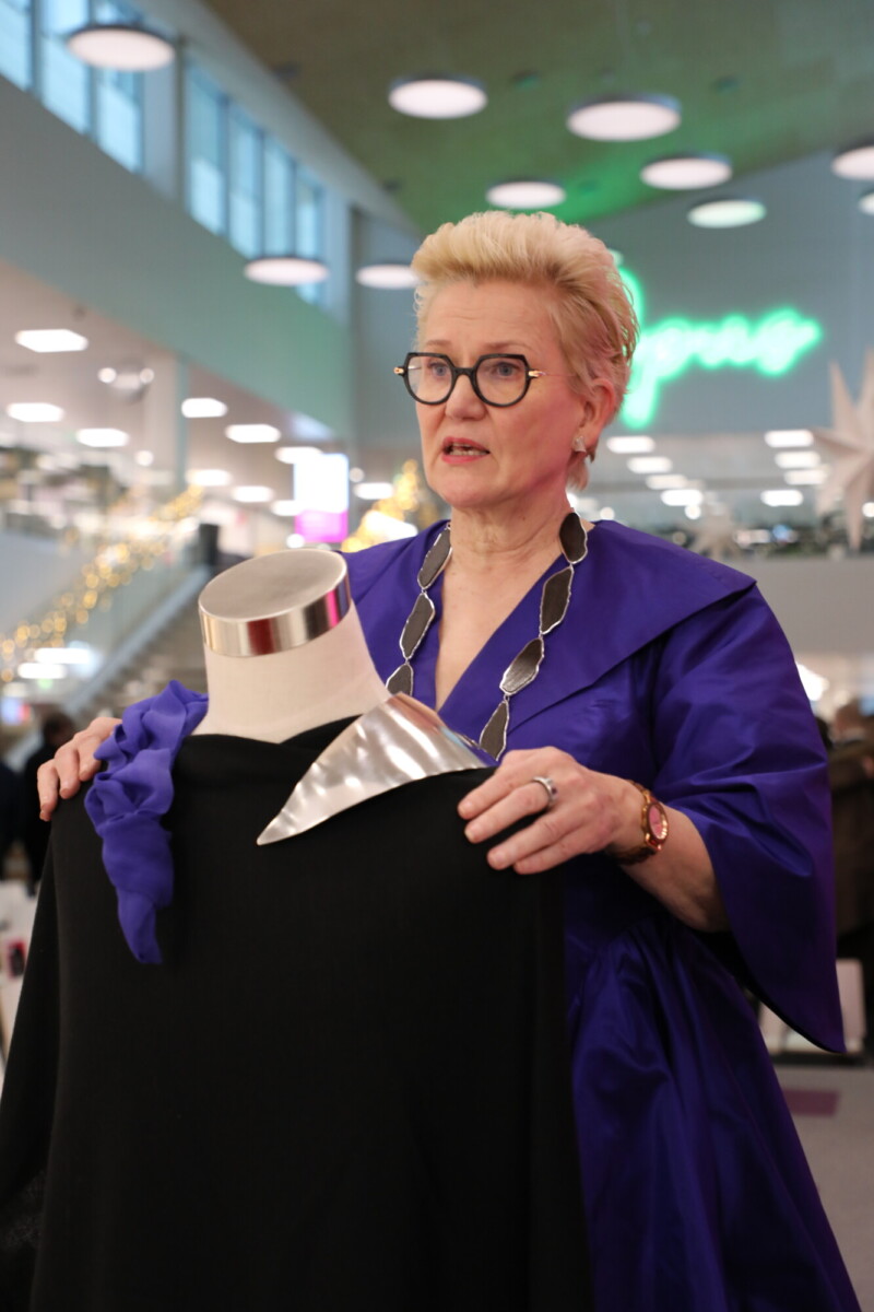 Violettiin mekkoon ja silmälaseihin pukeutunut nainen seisoo sisätiloissa ja pitelee mustaa vaatetta mallinuken päällä. Hän näyttää selittävän jotain, ja taustalla näkyy kirkkaita valoja ja koristeita.