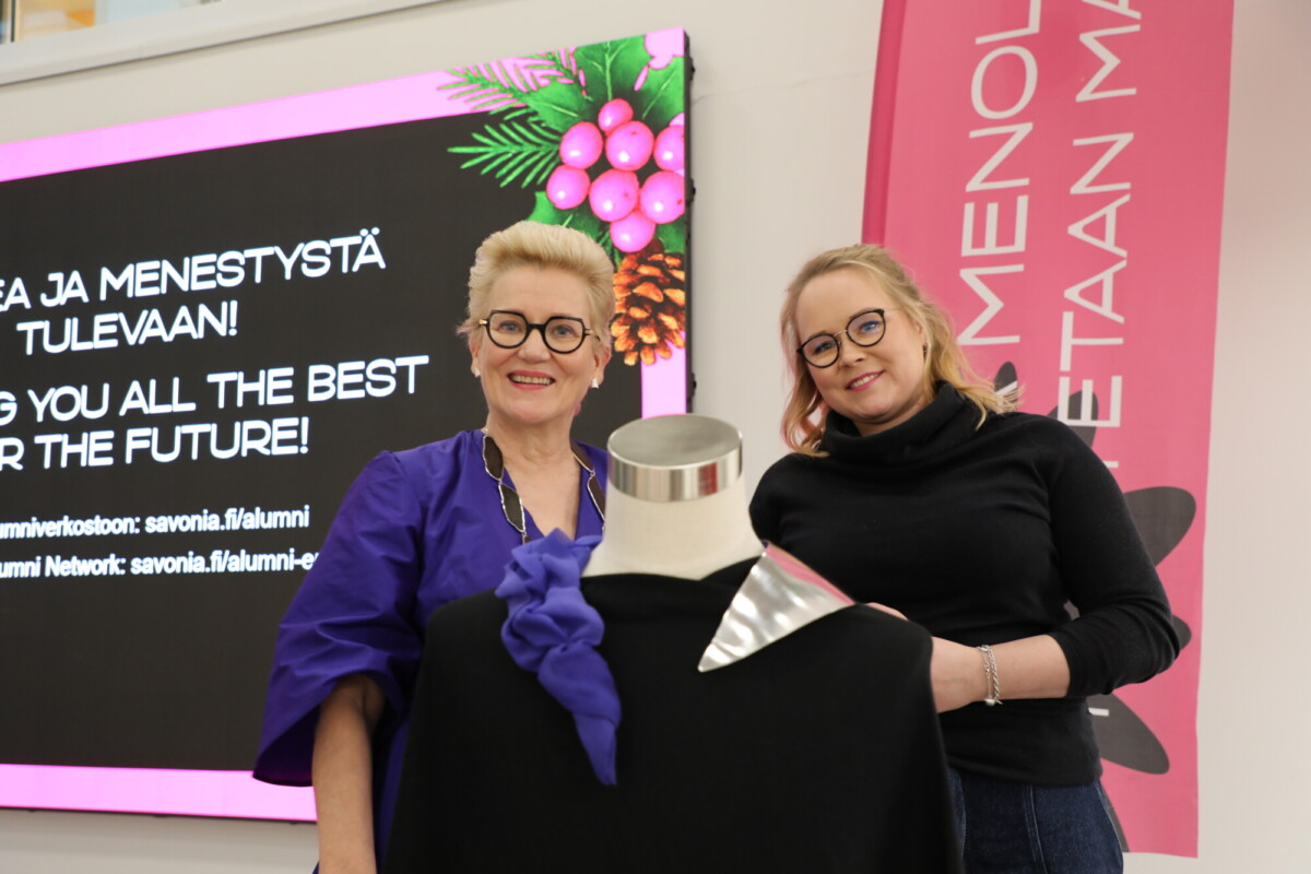 Kaksi naista seisoo hymyillen mallinuken vieressä, jolla on musta vaate ja hopeinen kaulus. Heidän takanaan on ruutu, jossa on suomenkielistä ja englanninkielistä tekstiä ja vasemmassa yläkulmassa juhlava kukkagrafiikka.