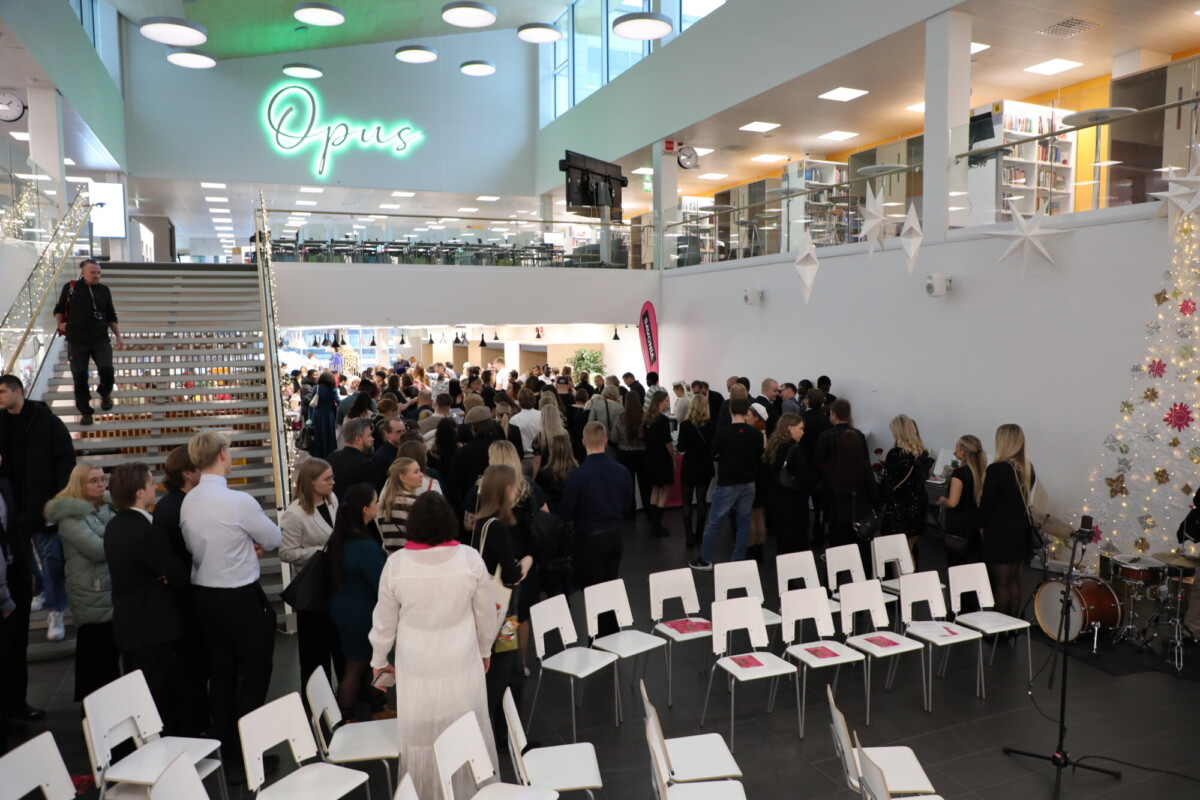 Suuri joukko ihmisiä kokoontuu moderniin, valoisaan rakennukseen, jossa on lasiseinä, portaikko ja valkoisia tuoleja. Näkyvissä on koristeltu joulukuusi ja soittimia. Neonkyltti Opus loistaa väkijoukon yllä.