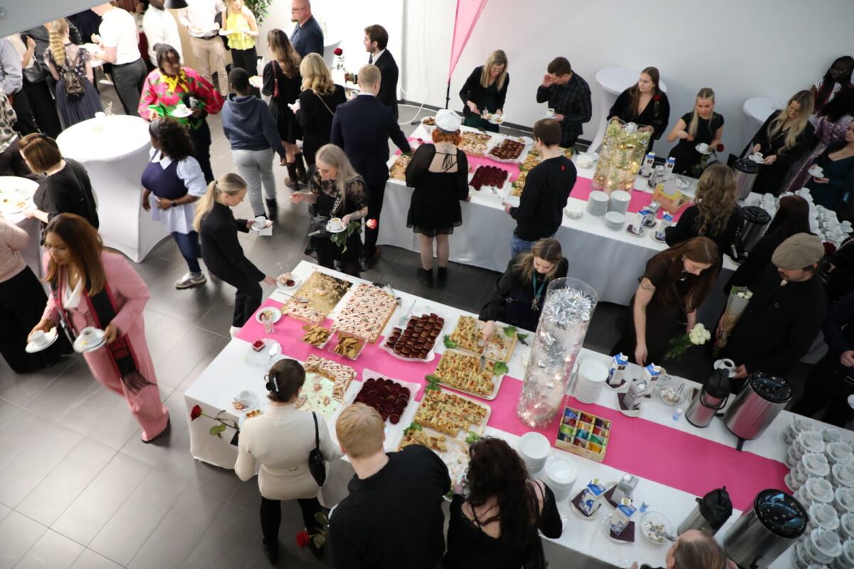 Joukko ihmisiä kerääntyy pöytien ympärille, joissa on erilaisia ruokia, juomia ja jälkiruokia, mikä näyttää olevan sosiaalinen tapahtuma tai vastaanotto. Jotkut tarjoilevat itseään, kun taas toiset keskustelevat valoisassa, modernissa tilassa.