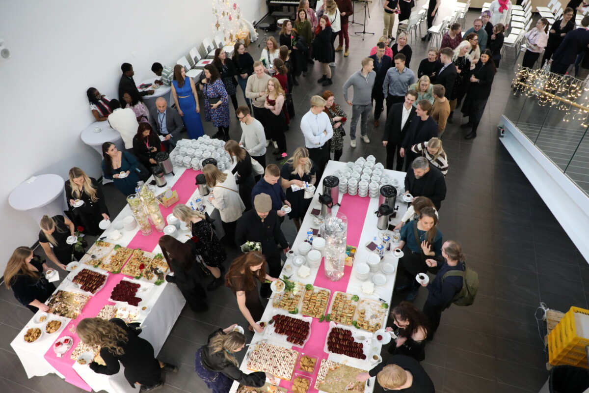 Ylhäältä näkymä suuresta ihmisryhmästä, joka on kerääntynyt pöytien ympärille, joissa on erilaisia välipaloja, leivonnaisia ja juomia vaaleanpunaisella pöytäliinalla sosiaalisessa tapahtumassa valoisassa sisätilassa.