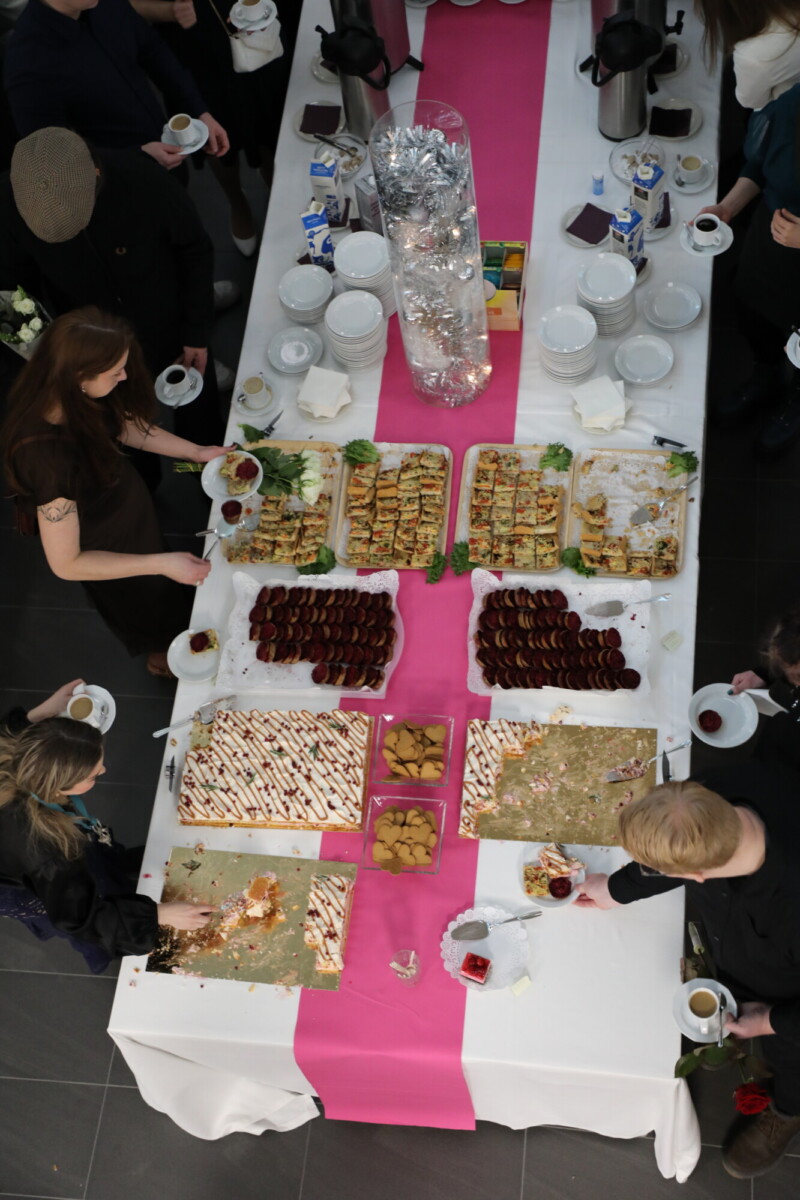 Ylhäältä katsottuna ihmiset tarjoilevat itselleen leivonnaisia, keksejä ja muita jälkiruokia pitkältä pöydältä, joka on peitetty vaaleanpunaisella ja valkoisella pöytäliinalla, lautasilla, kupeilla ja kahviannostelijoilla.