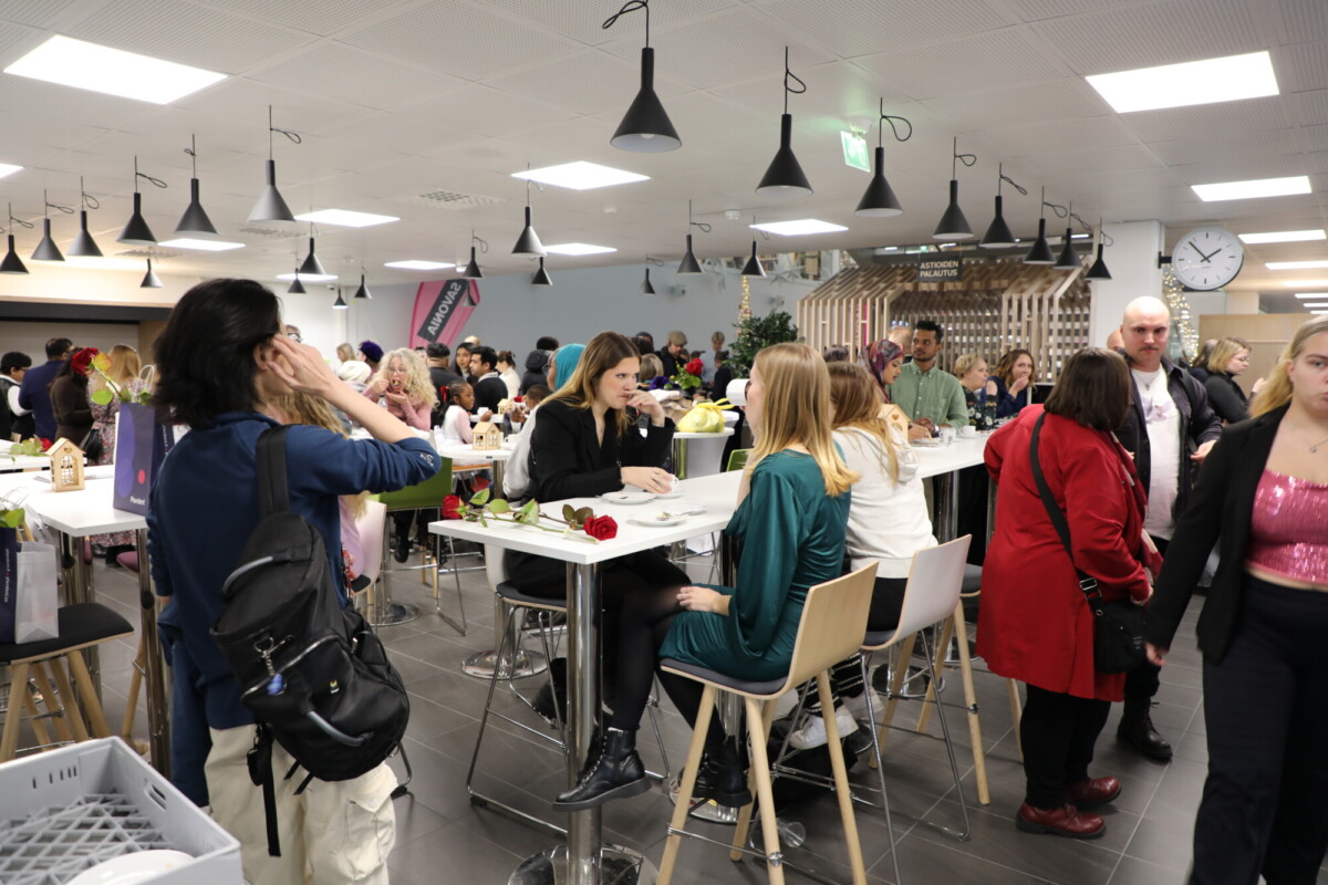 Kiireinen, moderni kahvila täynnä ihmisiä, jotka syövät, keskustelevat ja seisovat. Jotkut istuvat korkeissa pöydissä, toiset kävelevät tai seurustelevat. Katosta roikkuu ylävaloja, ja taustalla näkyy suuri seinäkello.