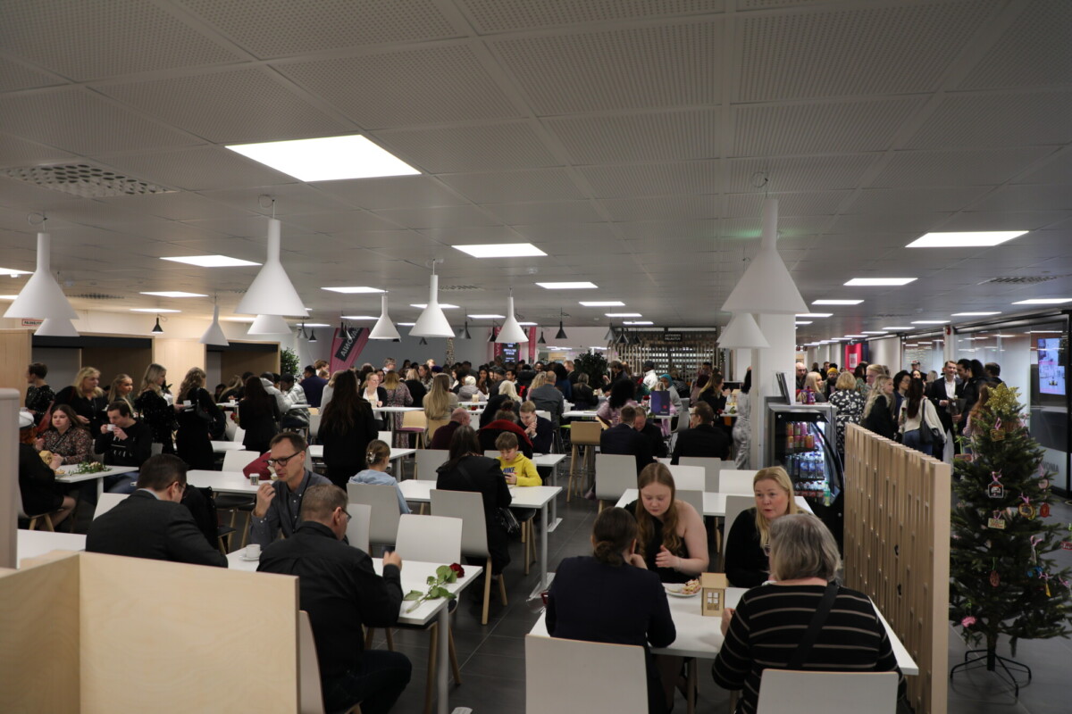 Ruuhkainen kahvila täynnä ihmisiä, jotka istuvat pöydissä syömässä ja puhumassa. Tilassa on moderni valkoinen valaistus, yksinkertaiset huonekalut ja pieni koristeltu joulukuusi sisäänkäynnin lähellä.