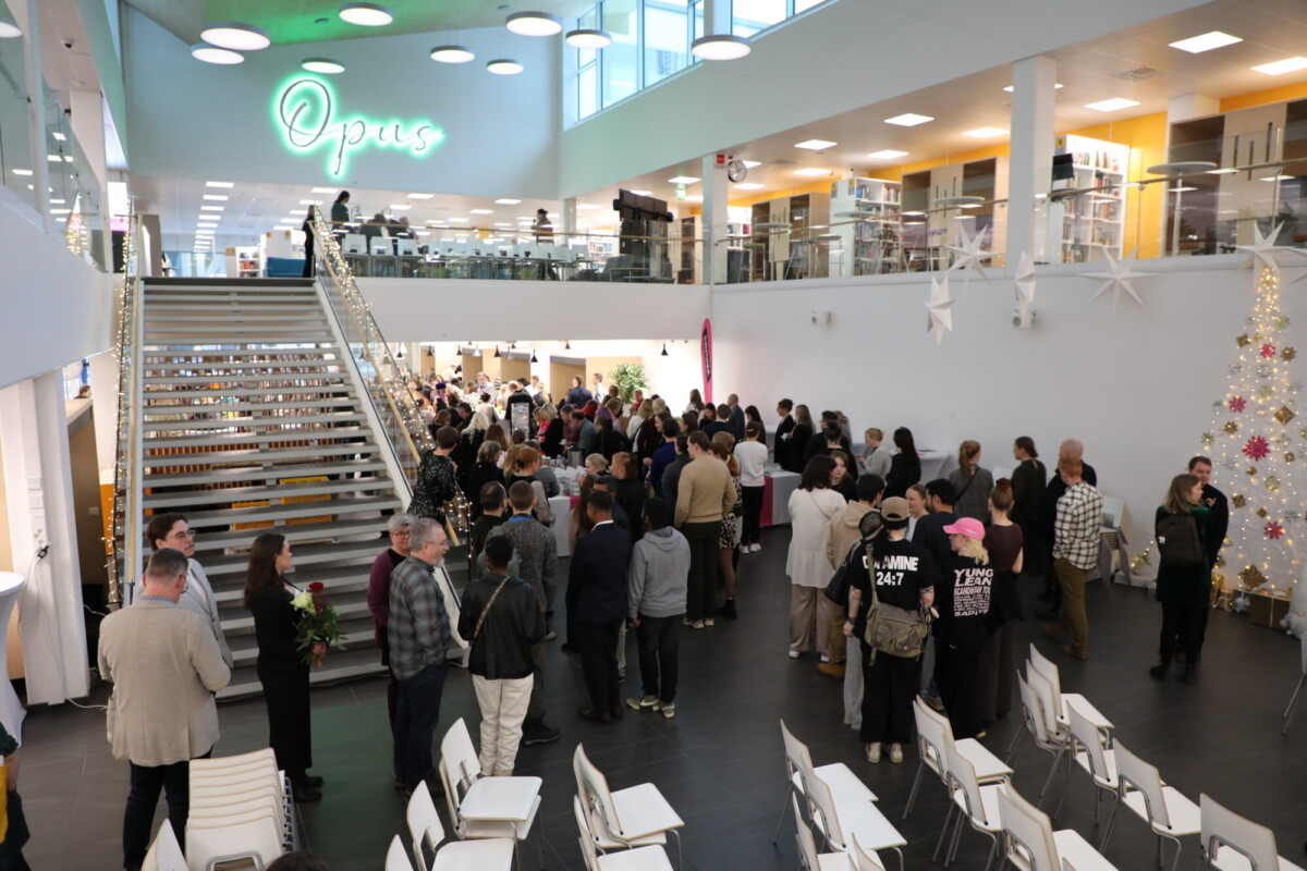 Suuri joukko ihmisiä kokoontuu tilavaan, moderniin rakennukseen, jossa on valkoiset seinät, koristeltu joulukuusi ja neonvalokyltti Opus. Osa ihmisistä seisoo jonossa portaikon vieressä, osa istuu tai sekoittuu joukkoon.