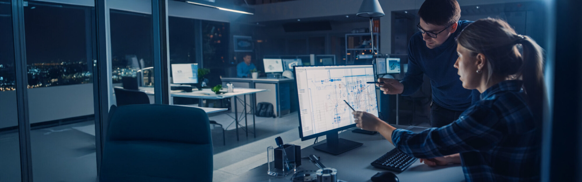 Kaksi ihmistä työskentelee myöhään toimistossa ja katselee suurta tietokoneruutua, jossa näkyy monimutkainen kaavio tai aikataulu. Toimisto on hämärästi valaistu, ja taustalla työskentelee muita työntekijöitä.