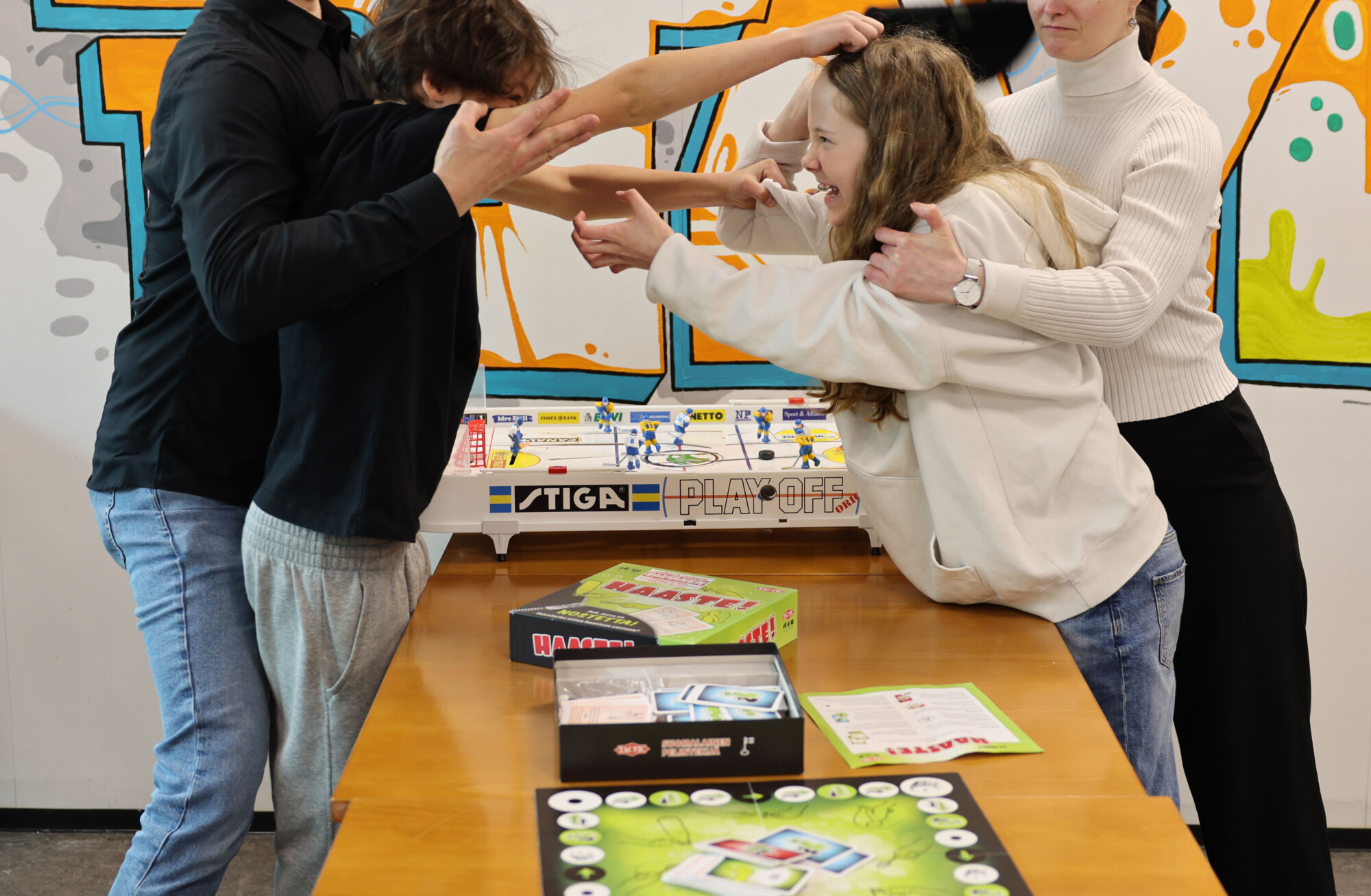 Neljä ihmistä seisoo pöydän ympärillä lautapelin kanssa, kaksi heistä riitelee ja osoittaa toisiaan sormilla, kun toinen yrittää erottaa heidät toisistaan. Taustalla on värikästä graffititaidetta.