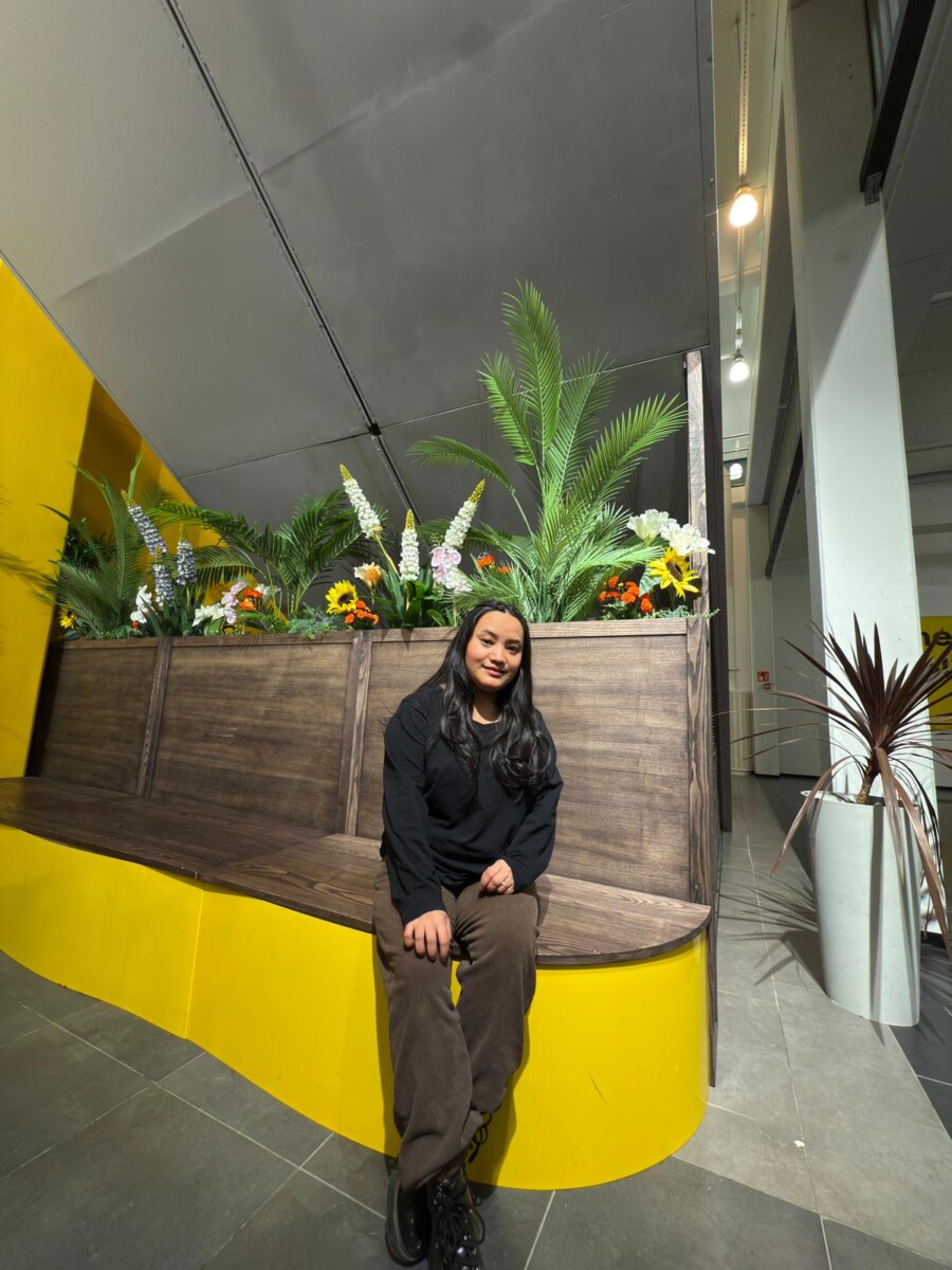 Nainen, jolla on pitkät tummat hiukset, pukeutunut mustaan toppiin ja ruskeisiin housuihin, istuu kaarevalla puupenkillä vihreitä kasveja ja värikkäitä kukkia sisältävien istutusastioiden edessä kirkkaasti valaistussa sisätilassa, jossa on keltaisia korostuksia.