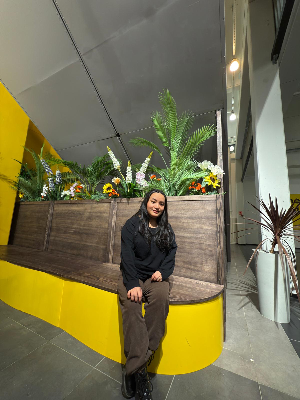 Nainen, jolla on pitkät tummat hiukset, pukeutunut mustaan toppiin ja ruskeisiin housuihin, istuu kaarevalla puupenkillä vihreitä kasveja ja värikkäitä kukkia sisältävien istutusastioiden edessä kirkkaasti valaistussa sisätilassa, jossa on keltaisia korostuksia.