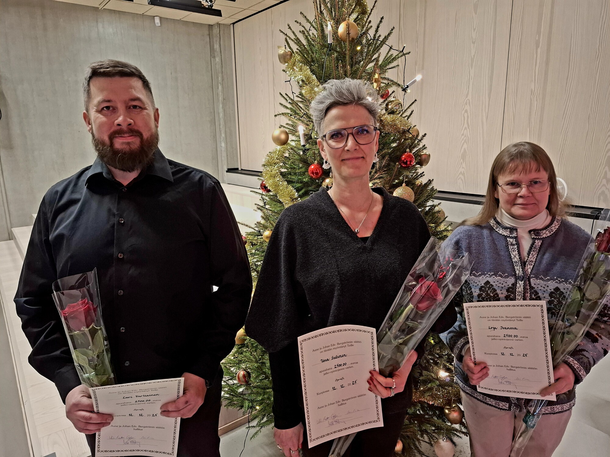 Kolme aikuista seisoo koristellun joulukuusen edessä, jokaisella kädessään todistus ja yksi punainen ruusu. He ovat pukeutuneet viralliseen tai puoliviralliseen asuun ja näyttävät poseeraavan tunnustus- tai palkintoseremoniaa varten.