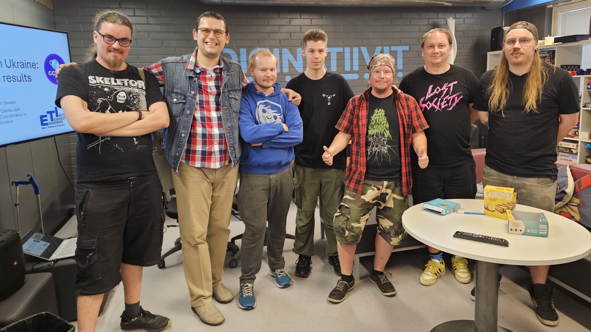 Seitsemän hengen ryhmä seisoo yhdessä modernissa huoneessa, jonka seinät ovat harmaat ja jossa on teollisuusputkia. He ovat pukeutuneet rennosti, jotkut heistä graafisissa t-paidoissa, ja hymyilevät kameralle. Taustalla on pöytiä, välipaloja ja valkokangas.