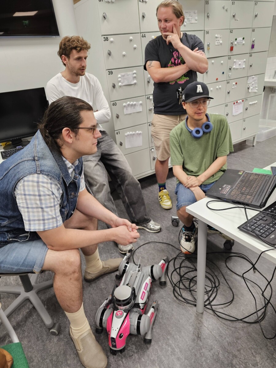 Neljä ihmistä on kerääntynyt pöydän ympärille kannettavan tietokoneen kanssa ja katselee tarkkaavaisesti näyttöä. Heidän edessään lattialla on vaaleanpunainen ja harmaa robottikoira. Heidän takanaan on kaappeja, joihin on kiinnitetty papereita ja laitteita.