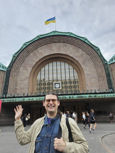 Hymyilevä silmälasipäinen henkilö vilkuttaa Helsingin päärautatieaseman edessä, kun sisäänkäynnin yläpuolella liehuu Ukrainan lippu ja taustalla kävelee ihmisiä.