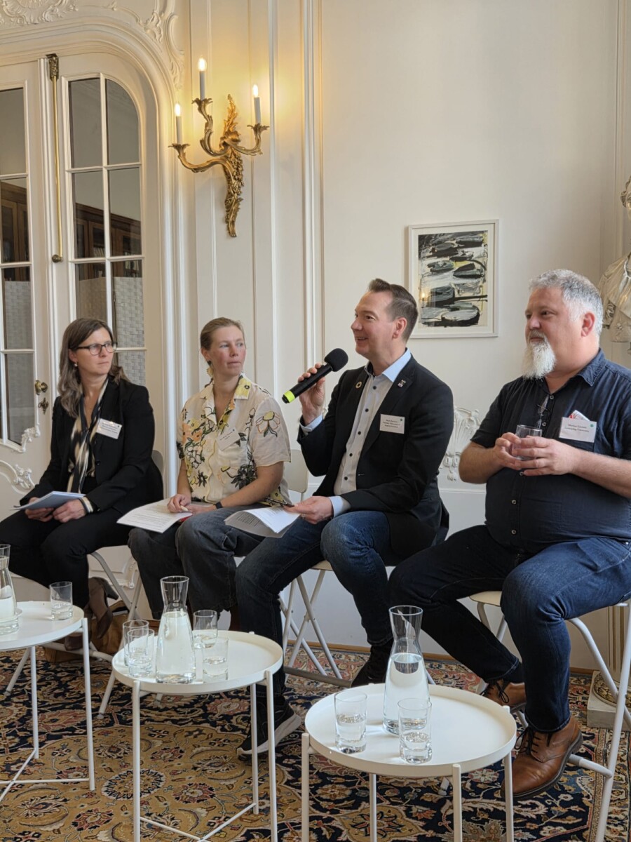 Neljä ihmistä istuu tuoleilla paneelikeskustelussa. Yksi mies puhuu mikrofoniin muiden kuunnellessa. He ovat sisätiloissa sisustetussa huoneessa, jossa on valkoiset seinät, pöydillä lasikannuja ja seinällä maalaus.