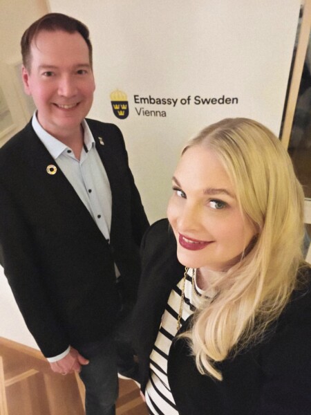 Mies ja nainen hymyilevät ja poseeraavat valokuvaa varten seinän edessä, jossa on Ruotsin Wienin suurlähetystön logo. Molemmat ovat pukeutuneet työasuun.
