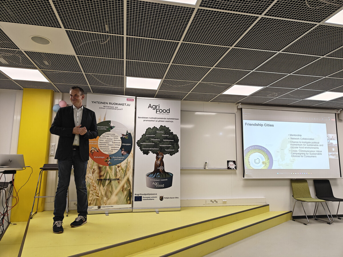 Mies seisoo pienellä lavalla pitämässä esitelmää. Hänen takanaan on maatalousgrafiikkaa sisältäviä banderolleja, ja hänen oikealla puolellaan näkyy dia, jonka otsikkona on Ystävyyskaupungit. Ympäristö on moderni kokoushuone.