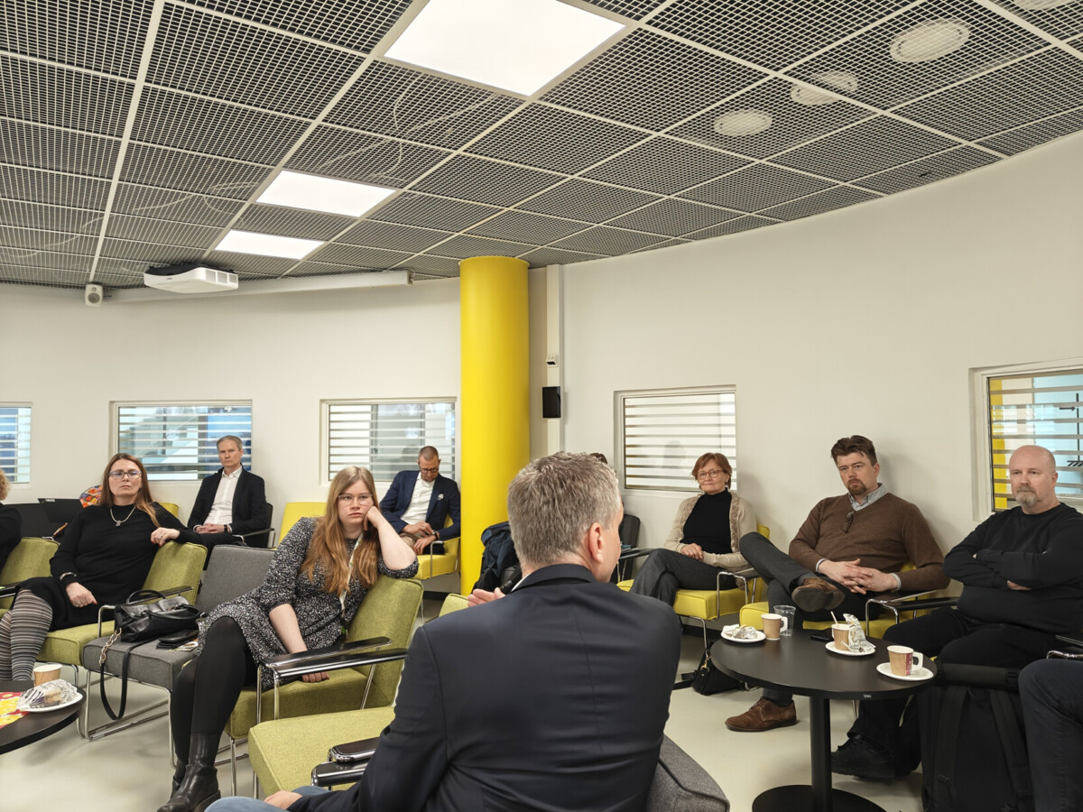 Joukko ihmisiä istuu modernissa kokoushuoneessa, jossa on keltaiset korostukset, ja kuuntelee tarkkaavaisesti. Pöydillä on kahvikuppeja ja muistikirjoja. Ilmapiiri on ammattimainen ja keskittynyt.