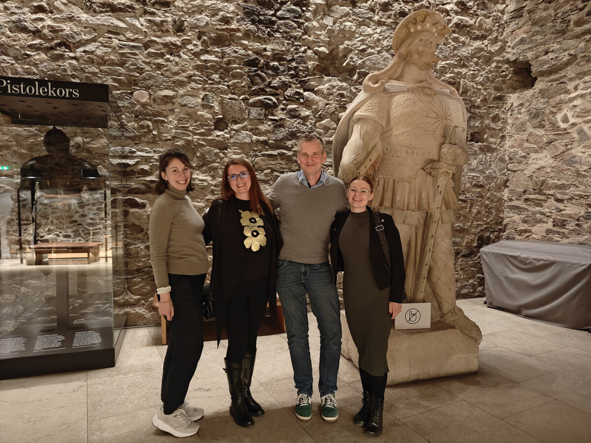 Neljä ihmistä seisoo hymyillen suuren kivisen soturipatsaan edessä kiviseinäisessä museossa. Kaksi naista on miehen kummallakin puolella, kaikki pukeutuneet rennosti. Taustalla näkyy vitriinejä.