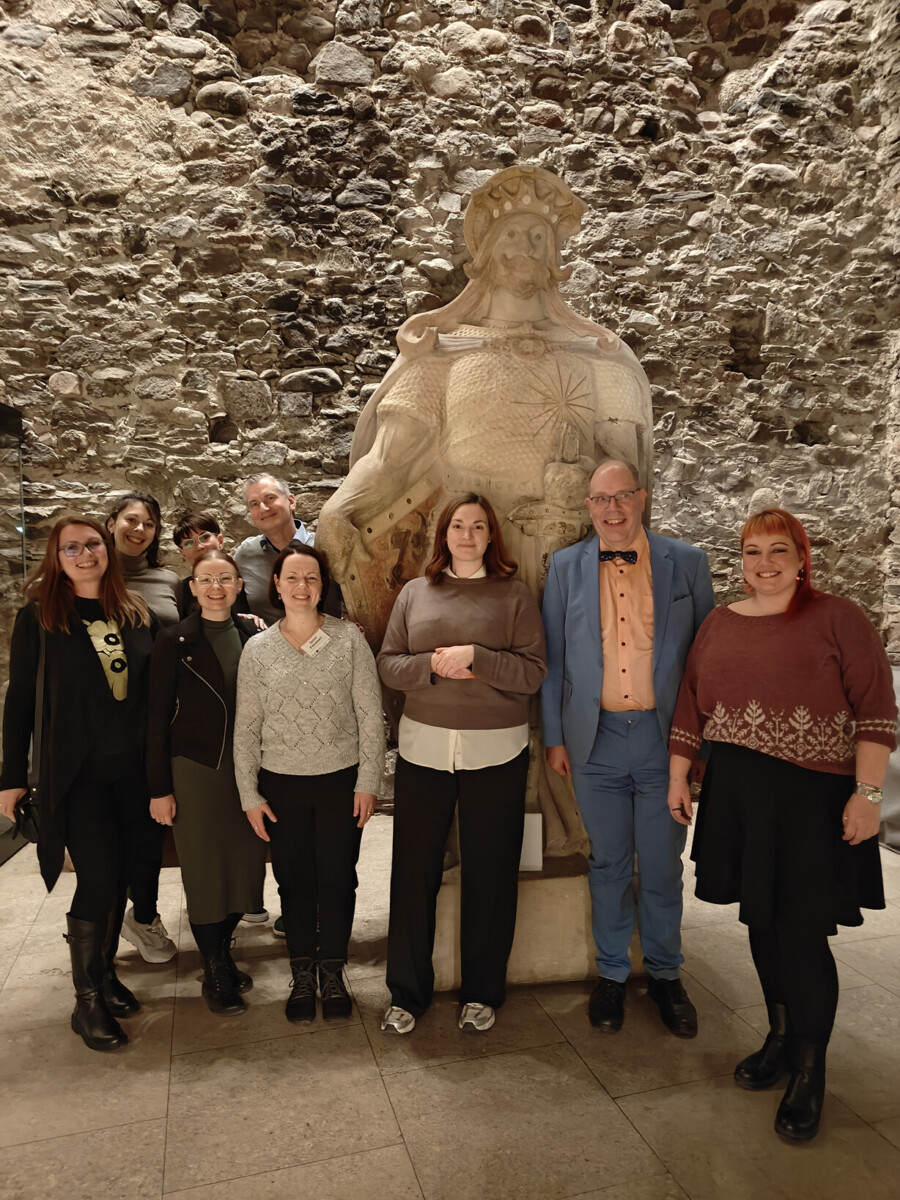 Kahdeksan hengen ryhmä poseeraa kruunattua hahmoa esittävän suuren kivipatsaan edessä museon tai historiallisen rakennuksen kiviseinää vasten.