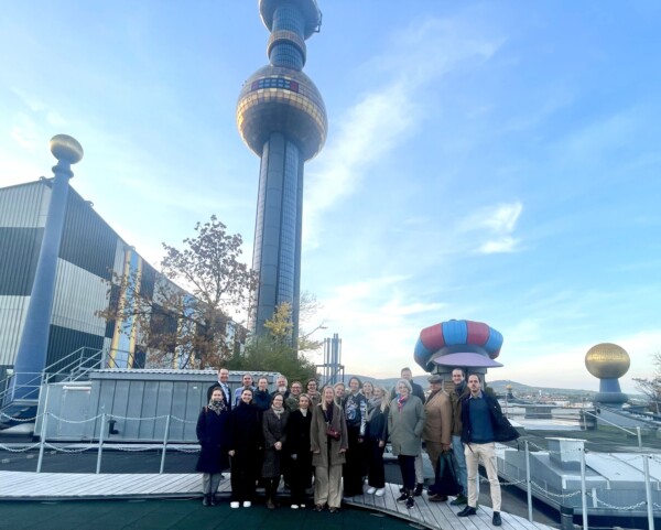 Joukko ihmisiä poseeraa yhdessä ulkona Wienin värikkään Spittelaun jätteenpolttolaitoksen edessä, jossa on tunnusomainen torni ja moderni, taiteellinen arkkitehtuuri sinisen taivaan alla.