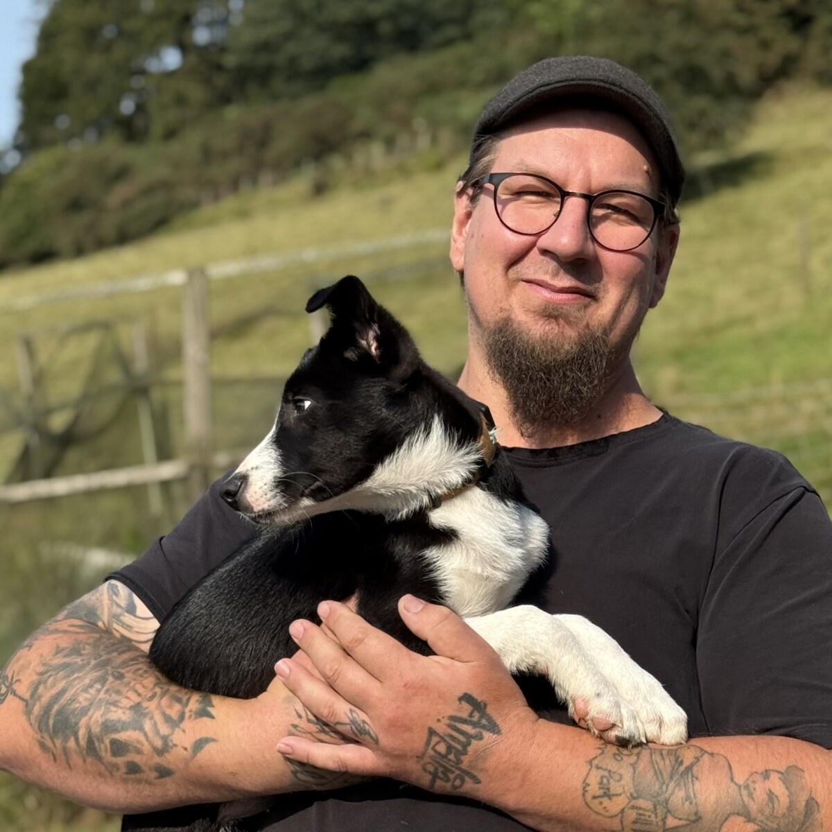 Silmälasipäinen ja tatuoitu henkilö hymyilee pitäessään mustavalkoista koiraa ulkona aurinkoisena päivänä, taustalla vihreää ruohoa ja puita.