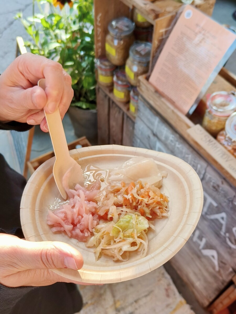 Henkilö pitelee puulautasta, jossa on kolmenlaisia marinoituja vihanneksia, ja käyttää puuhaarukkaa. Taustalla näkyy myyntikoju, jossa on purkkeja, ruokalista ja kukkia.