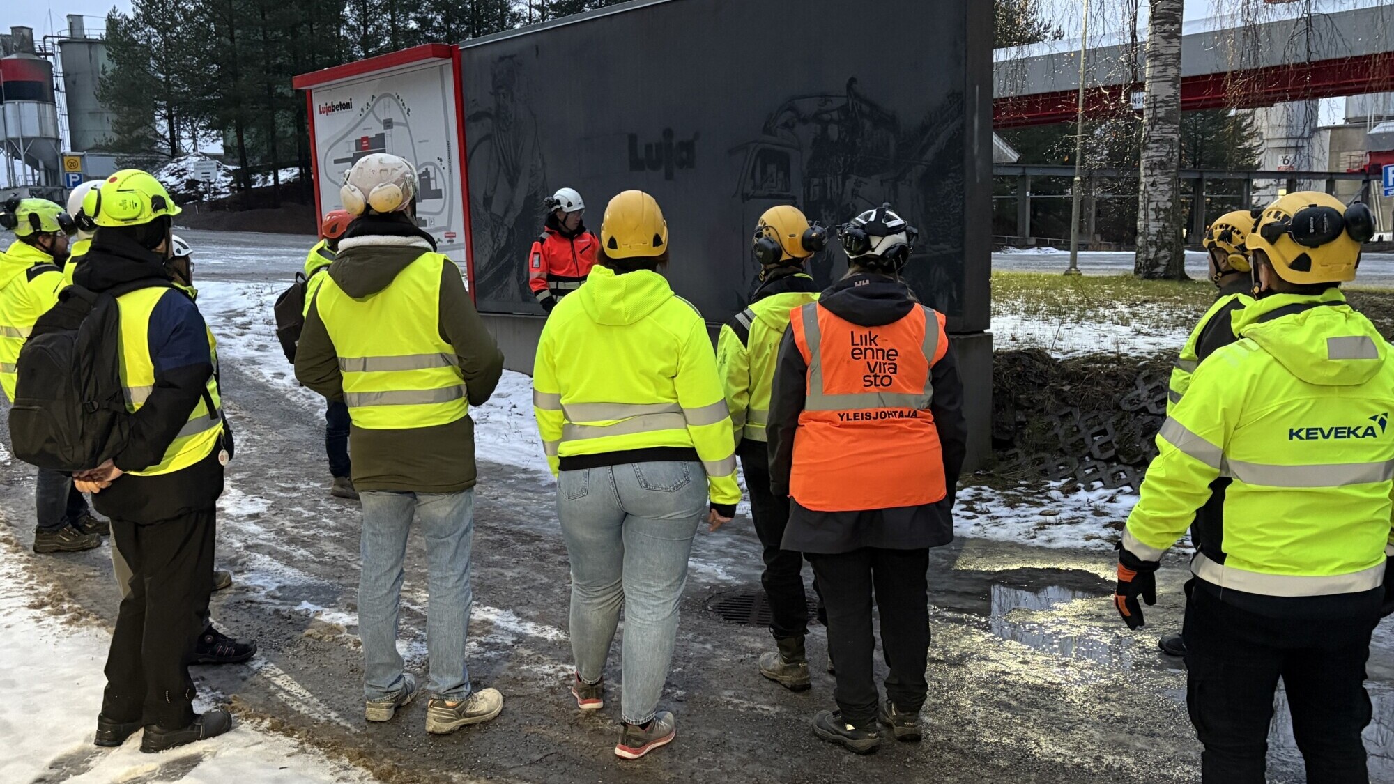 Joukko kypärä- ja näkösuojatakkeja käyttäviä ihmisiä kokoontuu ulkona lumisella pinnalla ja kuuntelee mustan kuorma-auton lähellä puhuvaa henkilöä. Taustalla näkyy puita ja rakennuksia.
