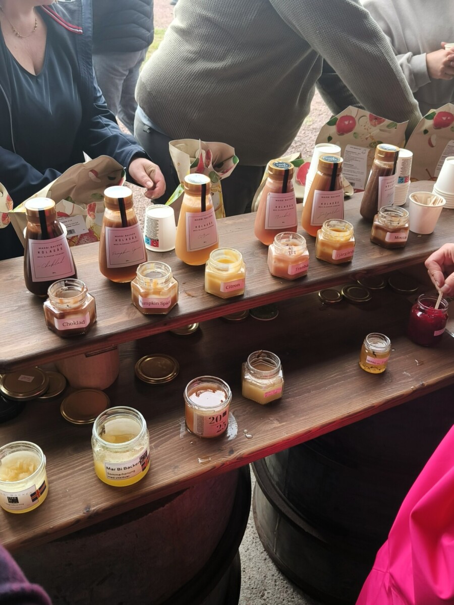 Hunajaa, mehua ja levitteitä sisältäviä purkkeja ja pulloja on esillä puuhyllyillä torikojulla. Ihmiset maistelevat ja täyttävät paperipusseja tuotteilla. Astioissa on näkyvissä erilaisia makuja ja etikettejä.