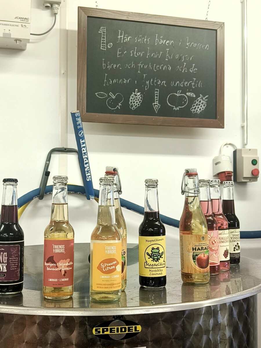 Metallisella tiskillä on esillä rivi erilaisia pullotettuja juomia. Niiden takana on liitutaulu, jossa on käsinkirjoitettua ruotsinkielistä tekstiä ja luonnoksia marjoista ja hedelmistä. Taustalla näkyy teollisuusputkia ja sähkölaitteita.