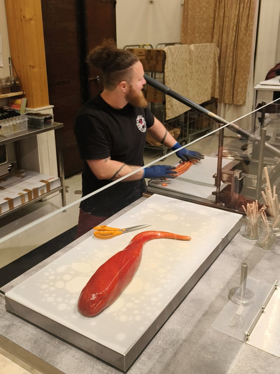 Lasinpuhaltaja, jolla on parta ja käsineet, työskentelee lasiseinän takana muotoillen lasisauvaa. Edessä olevalla pöydällä on oranssien lasityövälineiden vieressä suuri punainen kalaa muistuttava lasikappale.