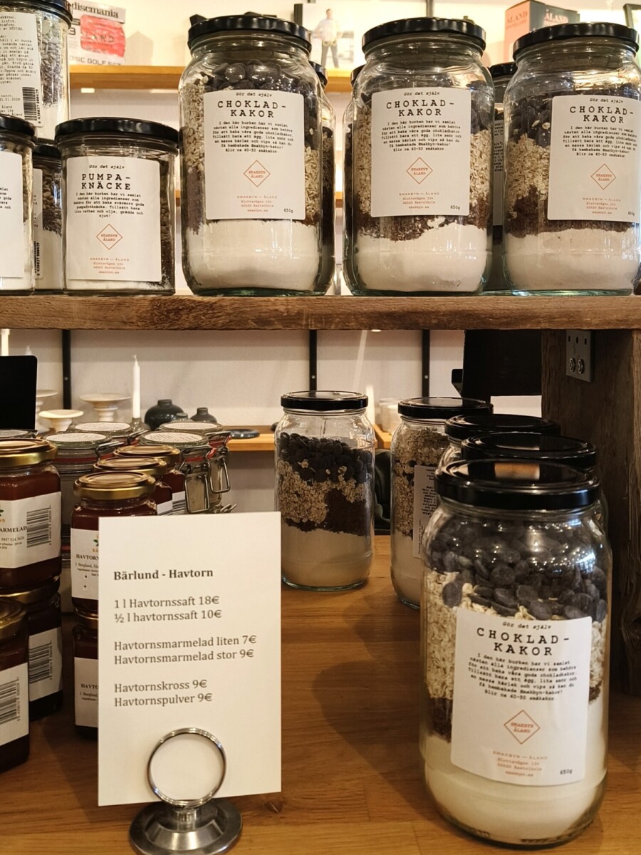 Puuhyllyillä on esillä lasipurkkeja, jotka on täytetty suklaakeksien ainesosilla. Lähistöllä on hillopurkkeja ja kyltti, jossa luetellaan erilaisten havtornituotteiden hinnat.