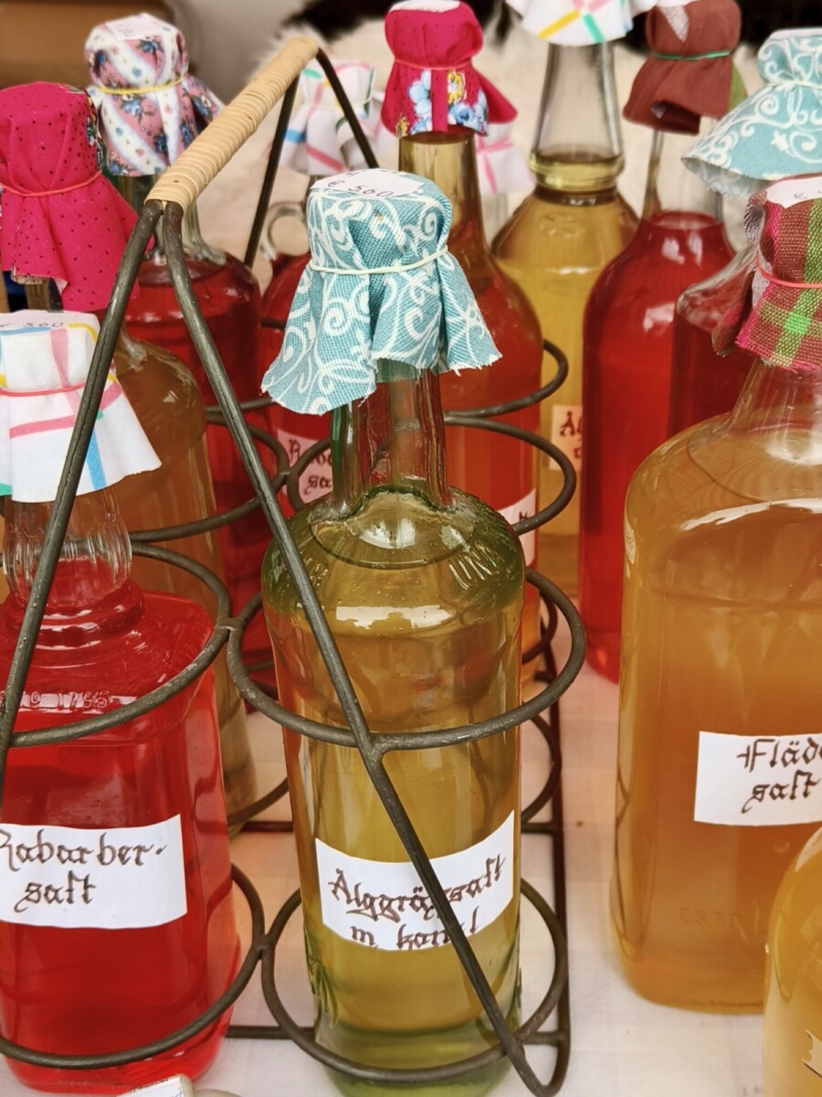 Useita värikkäillä kotitekoisilla mehuilla täytettyjä lasipulloja, jotka on päällystetty kuviollisella kankaalla ja merkitty saksaksi, on esillä vanhassa metallisessa pullotelineessä.