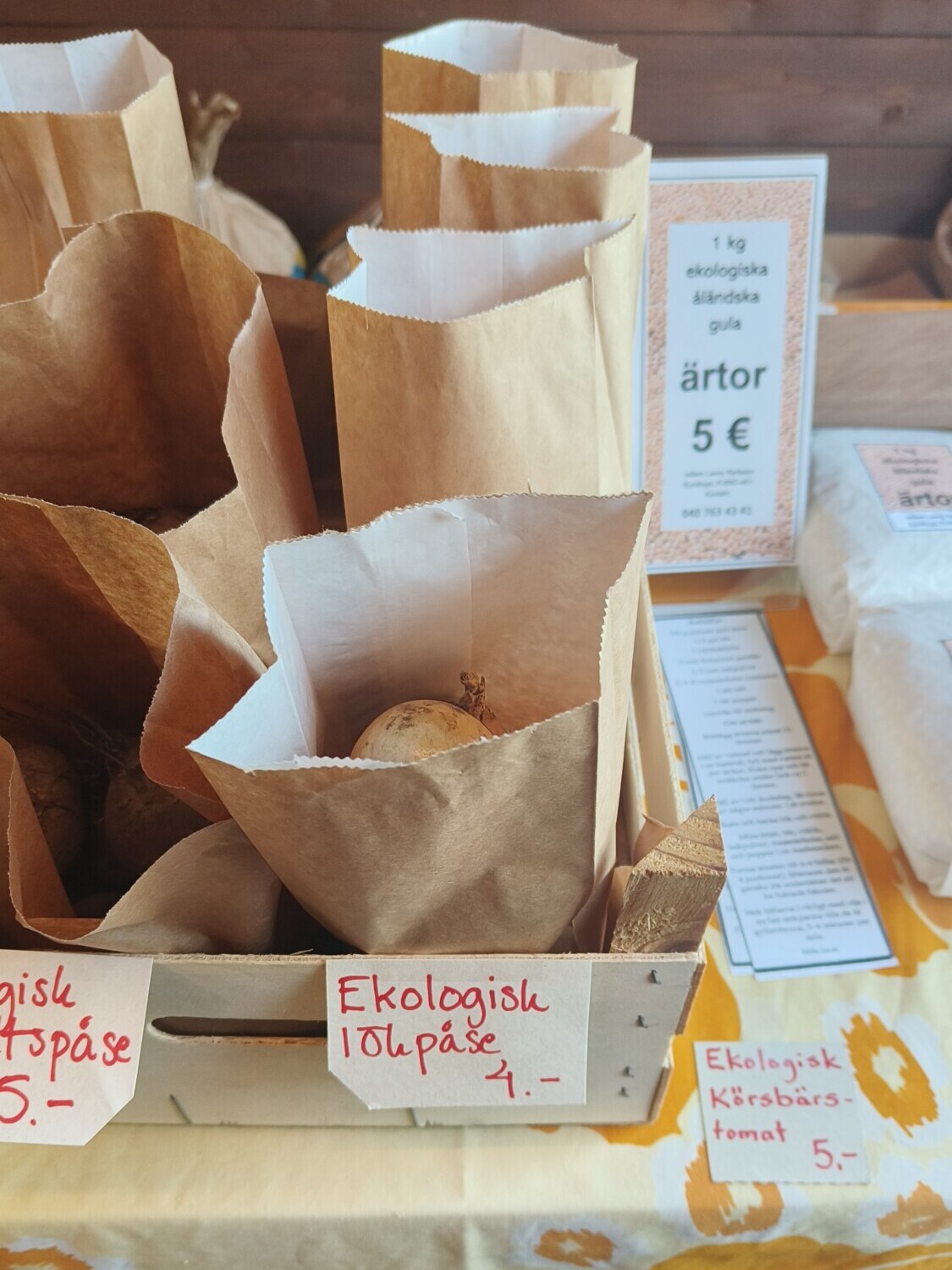 Luomuperunoilla täytetyt paperipussit istuvat puisessa laatikossa keltaisella kukkakuvioisella pöytäliinalla, ja taustalla on käsinkirjoitettuja ruotsinkielisiä hintakylttejä ja infokyltti.