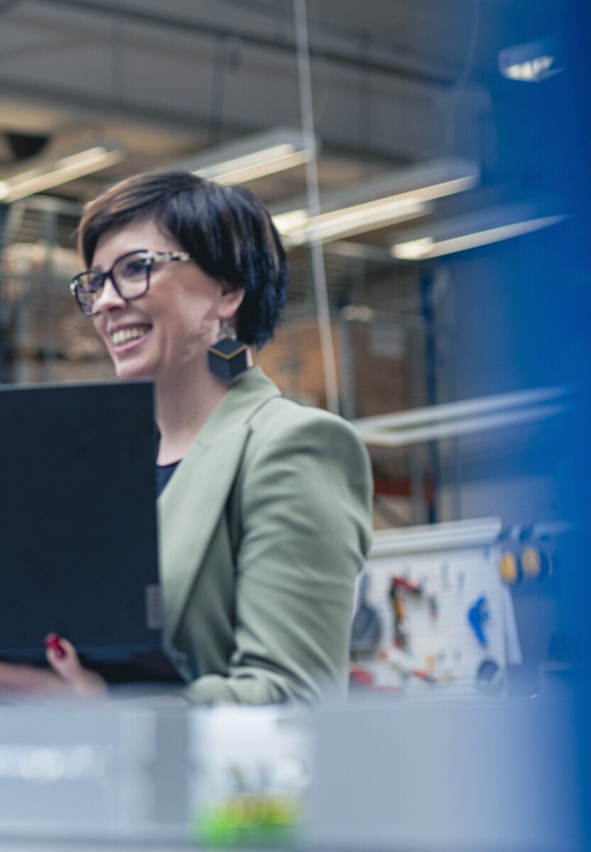 Lyhyet tummat hiukset ja silmälasit omaava, vihreään bleiseriin pukeutunut nainen hymyilee kannettavaa tietokonetta kädessään modernissa, teollisen näköisessä työtilassa.