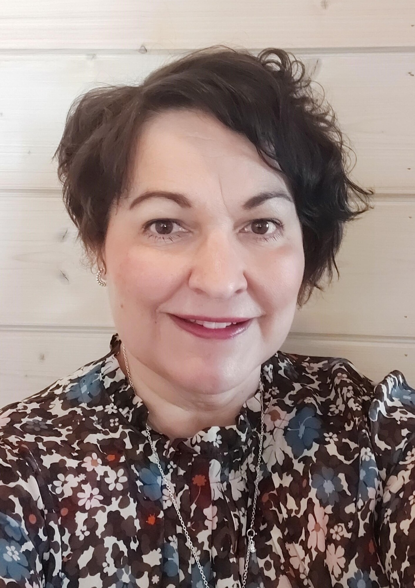 Nainen, jolla on lyhyet, laineikkaat tummat hiukset, hymyilee kameralle. Hänellä on yllään kukkakuvioinen pusero ja kaulakoru, ja hän poseeraa vaalean puuseinän edessä.