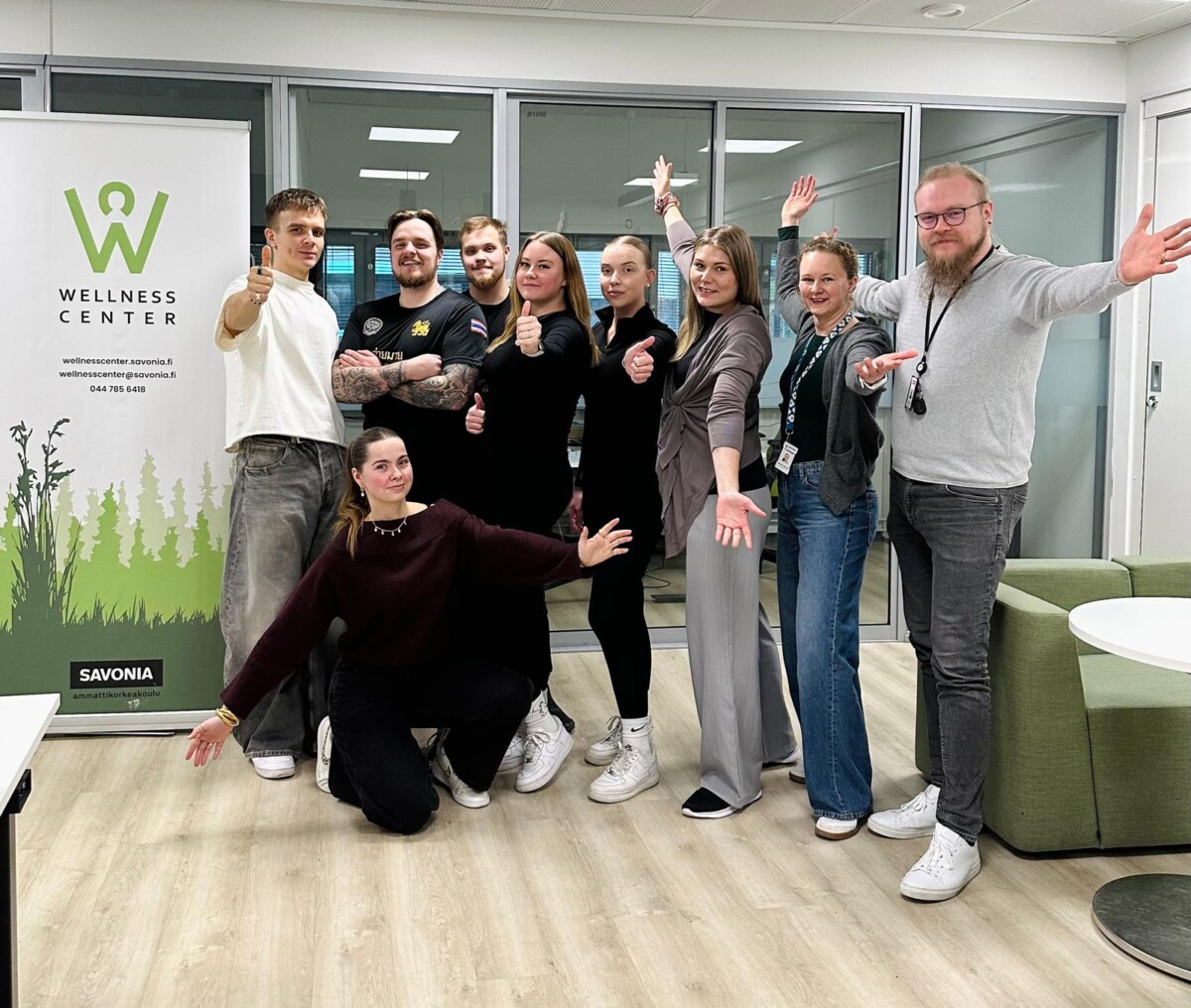 Yhdeksän ihmisen ryhmä poseeraa yhdessä, jotkut seisovat ja jotkut polvistuvat, hymyilevät ja nostavat peukaloita toimistotilassa banderollin vieressä, jossa lukee "Wellness Center Savonia".