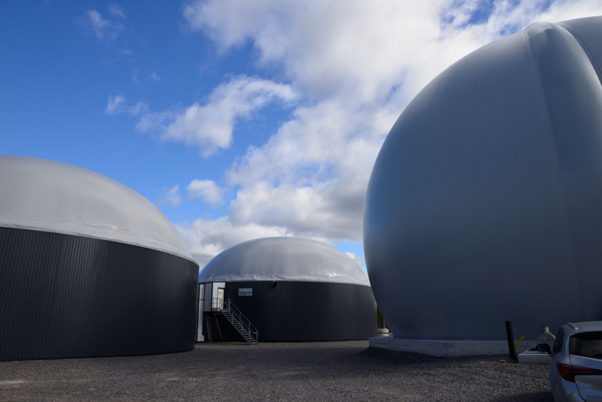 Kolme suurta, kupolimaista biokaasusäiliötä seisoo osittain pilvisen sinisen taivaan alla. Metalliportaat johtavat yhden säiliön ovelle, ja sorapeitteisen maan oikeassa reunassa näkyy auton osa.