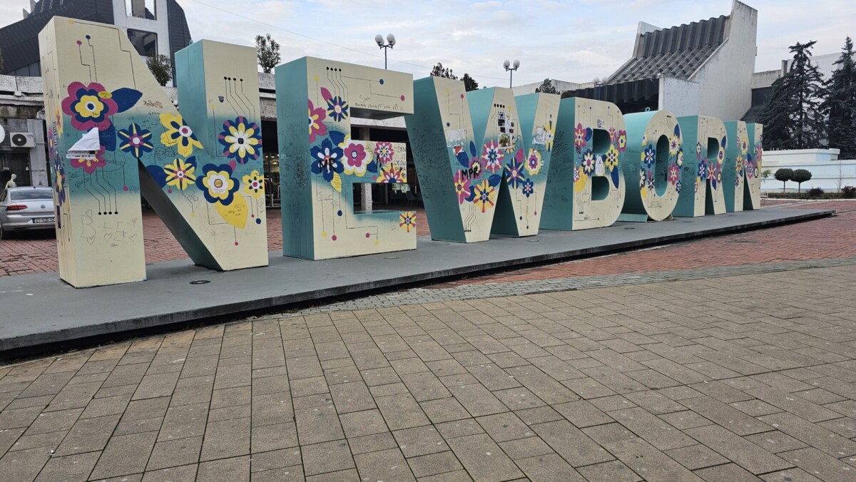 Suuret veistetyt kirjaimet, joissa lukee "NEWBORN", seisovat ulkona, värikkäillä kukkakuvioilla koristeltuina. Kyltti on korokkeella kaupungin aukiolla, ja taustalla näkyy rakennuksia ja puita.