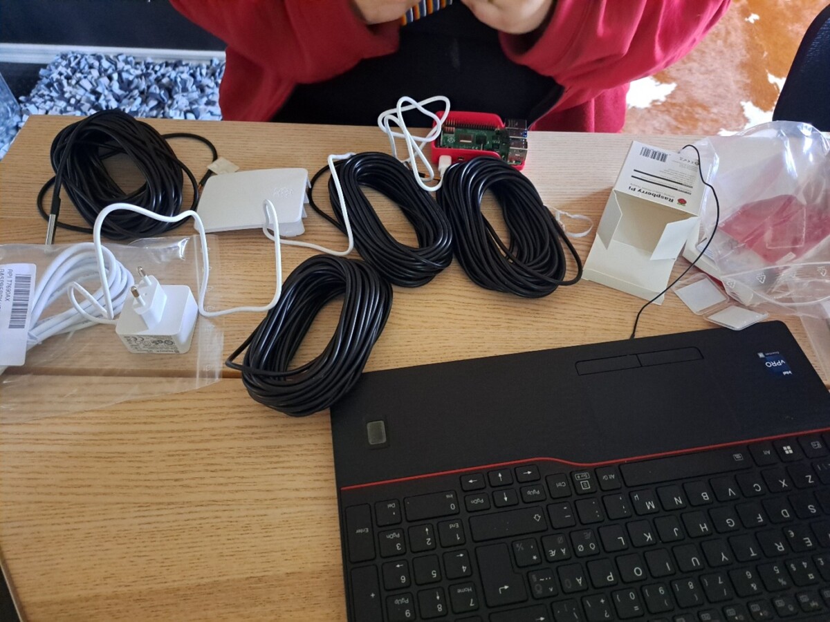 Punaiseen huppariin pukeutunut henkilö istuu pöydän ääressä, jossa on useita pitkiä mustia kaapeleita, sovittimia, virtapistoke, Raspberry Pi, avaamaton pakkaus ja musta kannettava tietokone. Kaapelit ja laitteet ovat levittäytyneet puupinnalle.