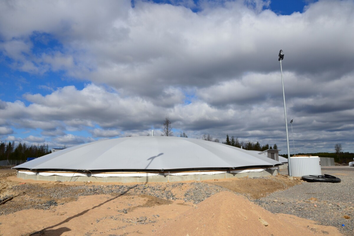 Suuri, kupolinmuotoinen, valkoisella päällysteellä varustettu rakennus seisoo soratontilla pilvisen taivaan alla, ja taustalla on valopylväitä ja harvahkoja puita.
