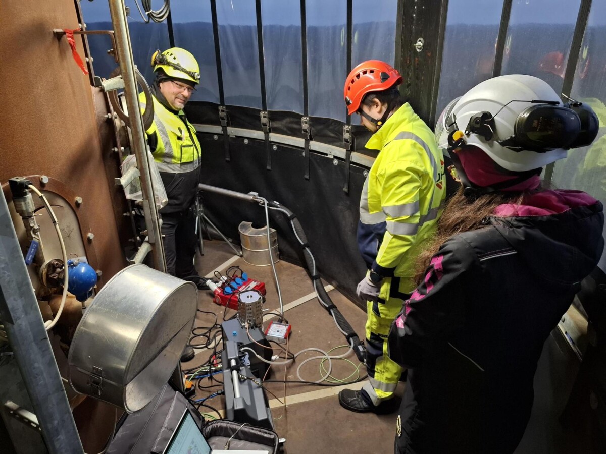 Kolme suojakypärä- ja suojavarusteisiin pukeutunutta henkilöä käyttää ja tarkkailee teknisiä laitteita ja kaapeleita metalliseinäisessä teollisuusympäristössä. Heidän ympärillään näkyy erilaisia välineitä ja koneita.