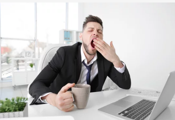 Pukumies istuu työpöydän ääressä kannettavan tietokoneen kanssa, muki kädessä ja haukottelemassa, ja vaikuttaa väsyneeltä valoisassa toimistoympäristössä.
