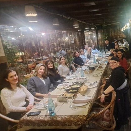 Joukko ihmisiä istuu pitkän, koristeellisesti koristellun ruokapöydän ympärillä viihtyisässä, lämpimästi valaistussa ravintolassa, jakaa aterian ja hymyilee kameralle.