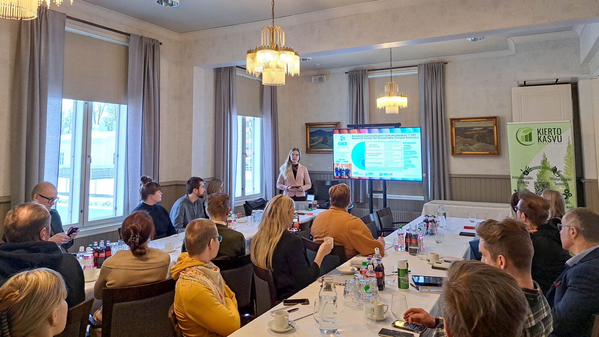 Joukko ihmisiä istuu suuren pöydän ympärillä kokoushuoneessa ja kuuntelee esittelijää, joka seisoo värikkään esityksen näyttävän näytön ääressä. Pöydällä on pulloja, papereita ja laseja. Ikkunoista virtaa valoa sisään.