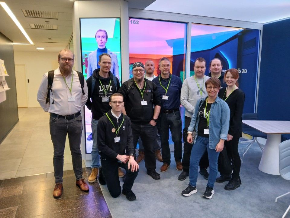 Kymmenpäinen ryhmä ihmisiä, joilla on kaulanauhat, poseeraa valokuvaa varten modernissa toimistossa, jossa on värikäs valaistus ja suuri digitaalinen näyttö taustalla. Jotkut seisovat ja toiset ovat hieman kyyryssä.