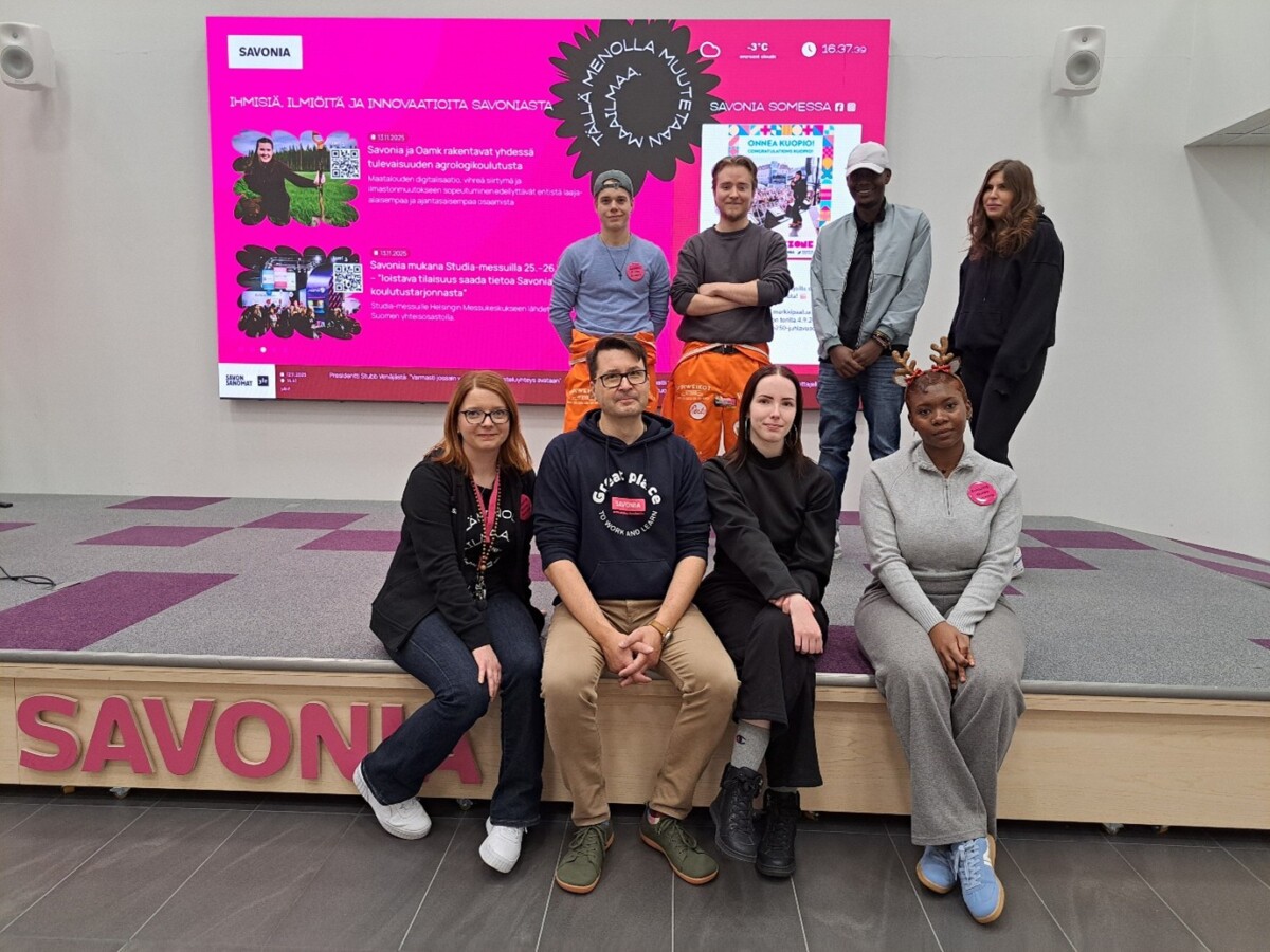 Kahdeksan ihmistä poseeraa ryhmäkuvassa sisätiloissa lavalla vaaleanpunaisen valkokankaan edessä. Jotkut seisovat ja toiset istuvat. Sana SAVONIA näkyy näyttämön etupuolella.