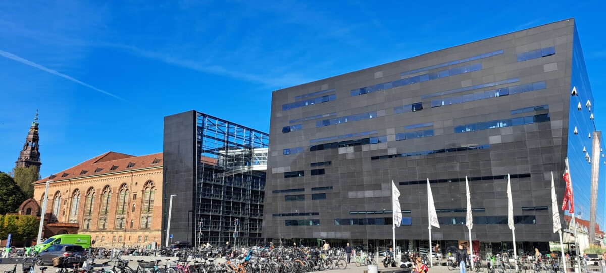 Moderni musta lasirakennus seisoo vanhemman tiilirakennuksen vieressä, jonka taustalla on korkea torni. Etualalle on pysäköity polkupyöriä kirkkaansinisen taivaan alla, ja kadun varrella on useita valkoisia lippuja.