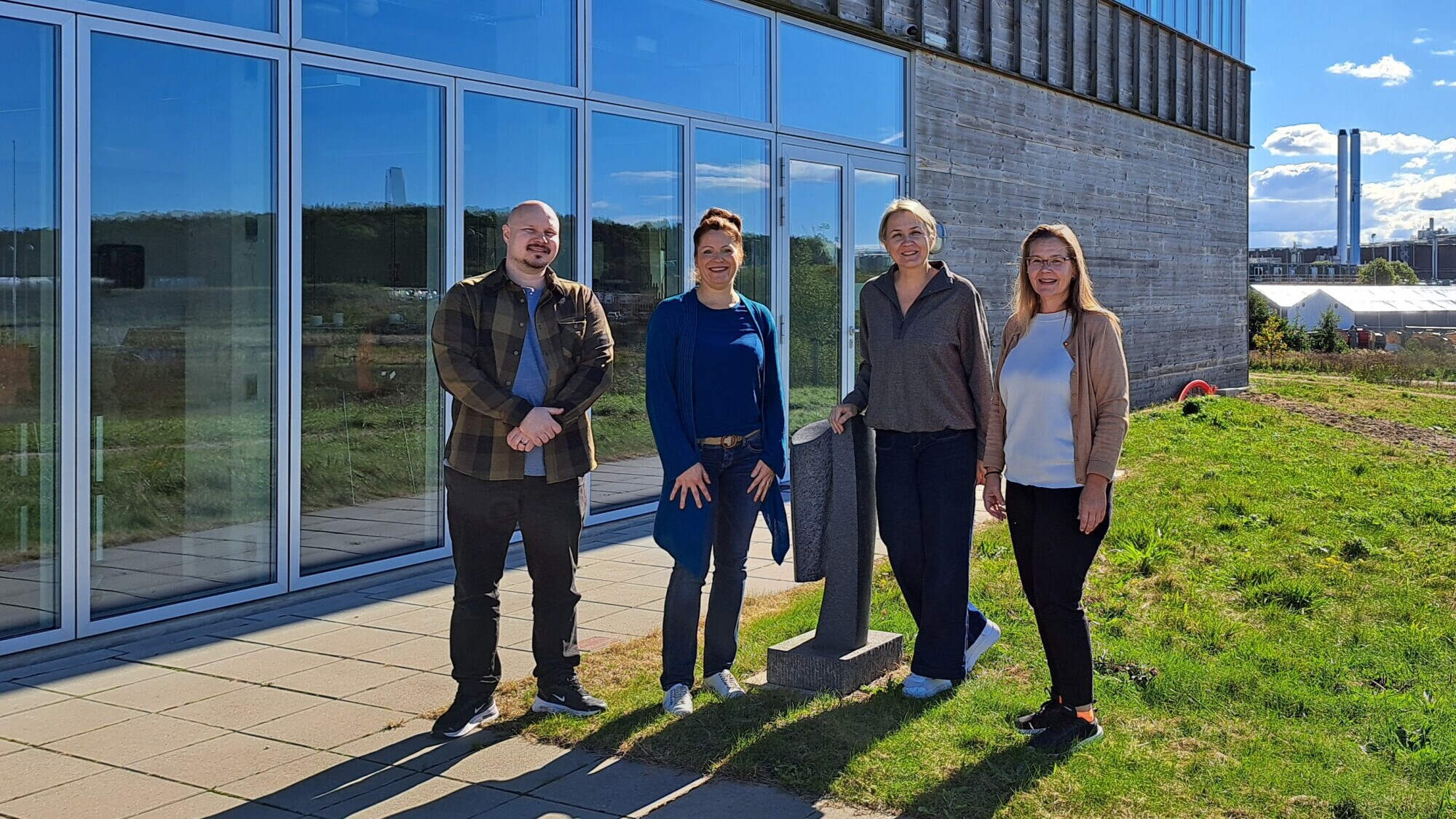 Neljä ihmistä seisoo modernin lasirakennuksen ulkopuolella aurinkoisena päivänä ja hymyilee kameralle. Ryhmä on päällystetyllä alueella, jonka läheisyydessä on nurmikkoa, ja taustalla näkyy teollisuuslaitos.