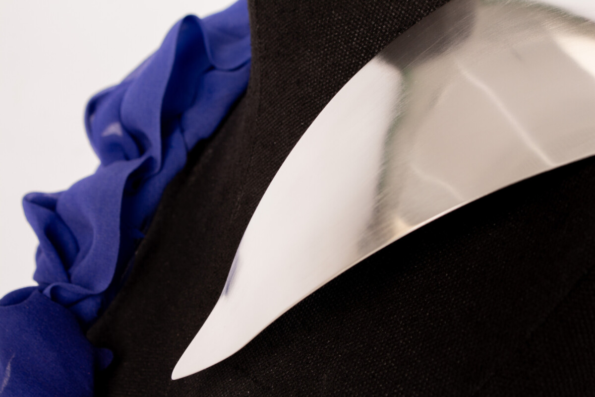 Lähikuva kiiltävästä, kaarevasta metallisesta lisävarusteesta mustalla kankaalla, jossa on koristeellinen sininen röyhelö, mahdollisesti osa vaatetta tai korua.